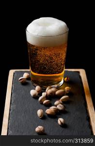 Glass of lager beer with pistachios nuts on stone board on black background.