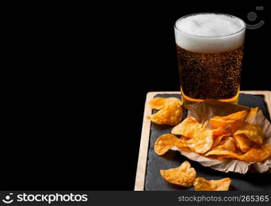 Glass of lager beer with potato crisps snack on stone board on black background.