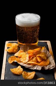 Glass of lager beer with potato crisps snack on stone board on black background.