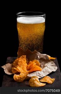 Glass of lager beer with potato crisps snack on vintage wooden board on black background.