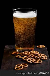 Glass of lager beer with pretzel snack on vintage wooden board on black.