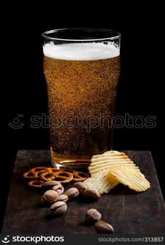 Glass of lager beer with snack on vintage wooden board on black. Pistachios and pretzel with potato crisps