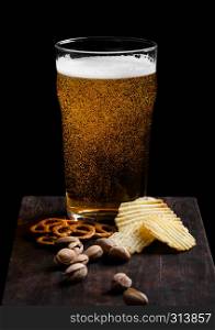 Glass of lager beer with snack on vintage wooden board on black. Pistachios and pretzel with potato crisps