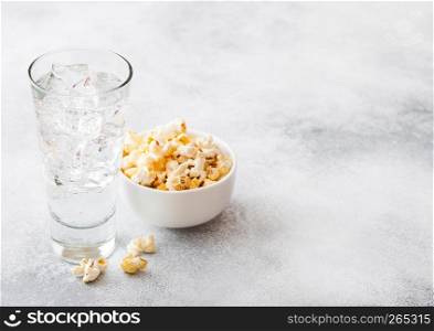 Glass of lemonade soda drink with ice cubes and whitel bowl of popcorn snack on stone kitchen background.