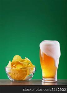 Glass of light beer with leaky foam and a dish of chips on a green background. Glass of light beer with leaky foam and dish of chips