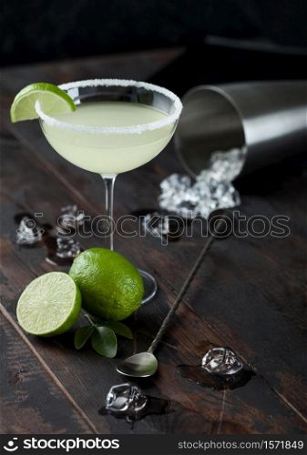 Glass of Margarita cocktail with fresh limes and bar spoon with ice cubes in shaker on wooden table background. Top view