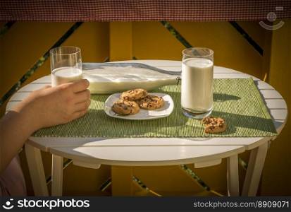 Glass of milk held in a woman s hand and a plate with cookies, on a white round table, in a warm sunny morning.