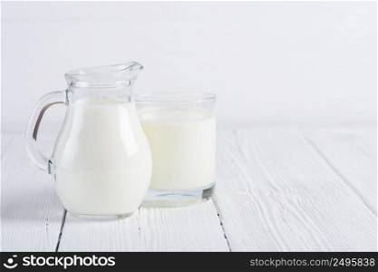 Glass of milk with small jug of milk on white wooden table still life