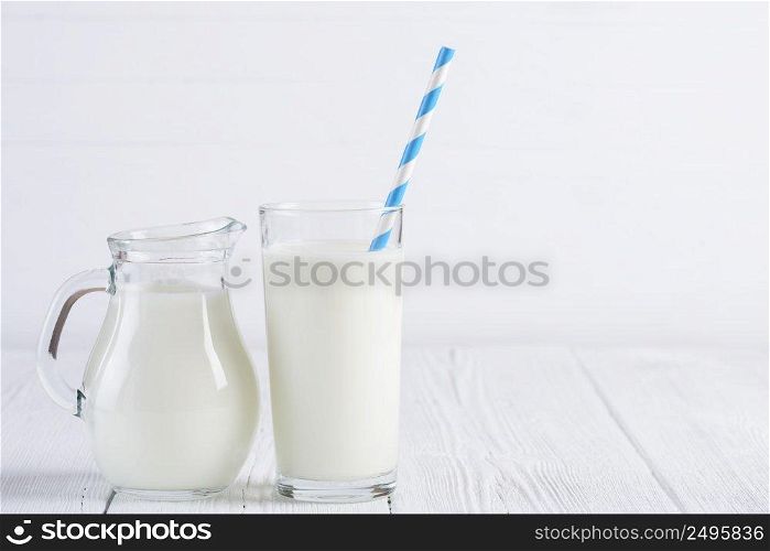Glass of milk with stripped blue paper straw and jug of milk on white wooden table