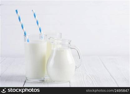 Glass of milk with stripped blue paper straw and jug of milk on white wooden table still life