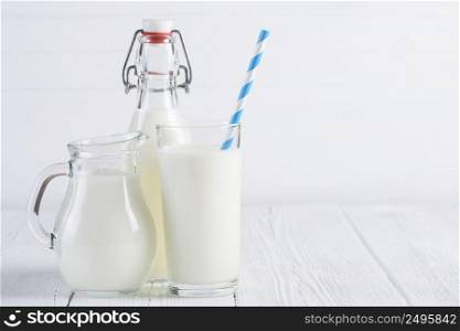 Glass of milk with stripped blue paper straw and jug with bottle of milk on white wooden table