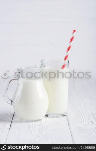 Glass of milk with stripped red paper straw and jug of milk on white wooden table