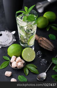 Glass of Mojito cocktail with ice cubes mint and lime on black board with spoon and fresh limes and cane sugar with wooden squeezer and black shaker with ice.Best summer cocktail