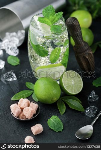 Glass of Mojito cocktail with ice cubes mint and lime on black board with spoon and fresh limes and cane sugar with wooden squeezer and steel shaker with ice.Best summer cocktail