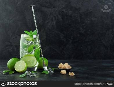 Glass of Mojito cocktail with ice cubes mint and lime on black board with spoon and fresh limes with cane sugar. Best summer cocktail. Space for text