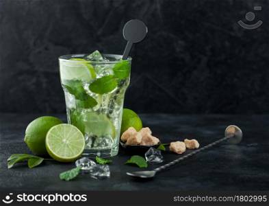 Glass of Mojito cocktail with ice cubes mint and lime on black board with spoon and fresh limes with cane sugar. Best summer cocktail