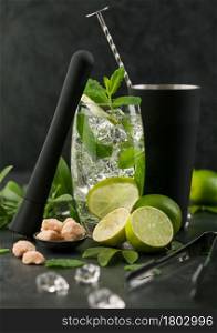 Glass of Mojito cocktail with ice cubes mint and lime on black board with spoon in shake and black muddler and fresh limes with cane sugar. Summer party drink.