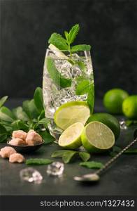 Glass of Mojito cocktail with ice cubes,mint and lime on black board with spoon and fresh limes with cane sugar.. Best party drink.