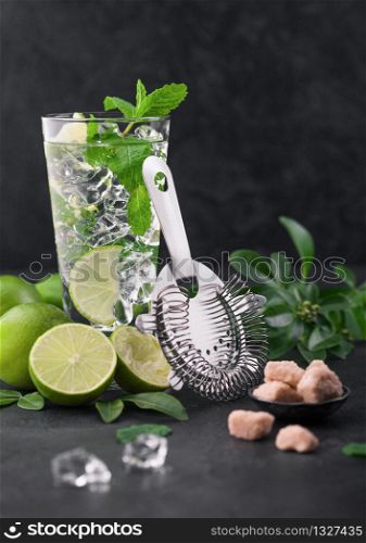 Glass of Mojito cocktail with ice cubes,mint and lime on black board with strainer and fresh limes with cane sugar. Best party drink.