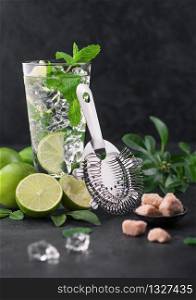Glass of Mojito cocktail with ice cubes,mint and lime on black board with strainer and fresh limes with cane sugar. Best party drink.