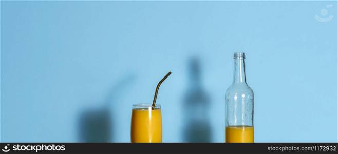 Glass of orange juice and empty bottle on blue background. Freshly squeezed orange juice in a glass and metal straw. Summer tropical drink banner.