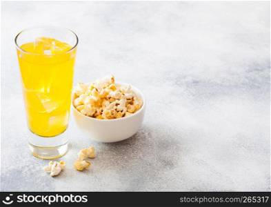 Glass of orange soda drink with ice cubes and whitel bowl of popcorn snack on stone kitchen background.