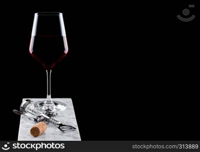 Glass of red wine on marble board with corkscrew opener and cork on black.