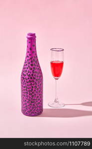 Glass of red wine with bottle painted hot pink with black spots and hard shadows on a pastel pink background, copy space.. Open bottle painted black spots with glass of red wine with shadows.