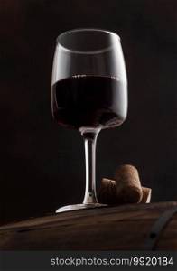 Glass of red wine with corks on top of wooden barrel on black background.