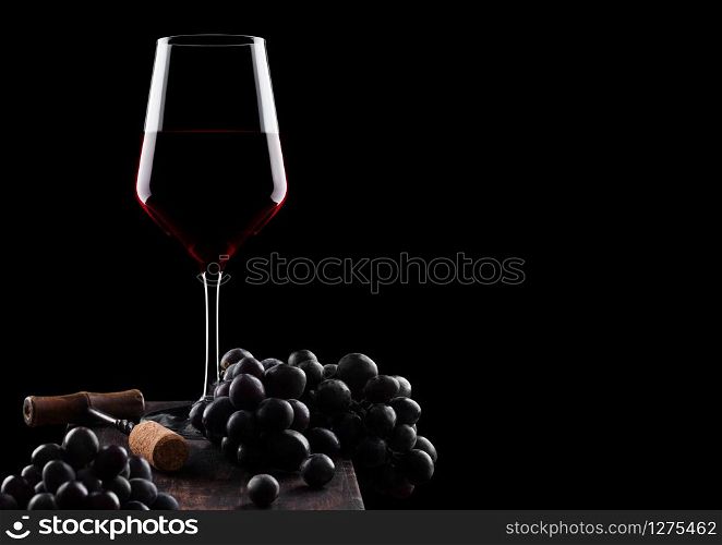 Glass of red wine with dark grapes and vintage corkscrew opener and cork on wooden board on black background.