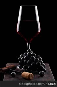 Glass of red wine with dark grapes and vintage corkscrew opener and cork on wooden board on black