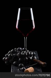 Glass of red wine with dark grapes and vintage corkscrew opener and cork on wooden board on black