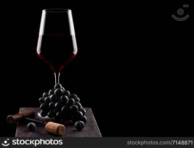 Glass of red wine with dark grapes and vintage corkscrew opener and cork on wooden board on black background.