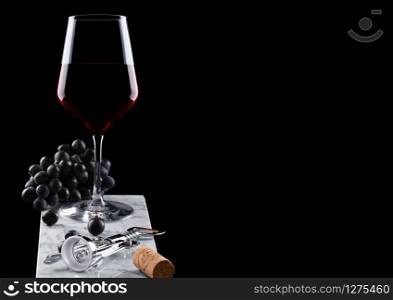 Glass of red wine with dark grapes on marble board with corkscrew opener and cork on black background.