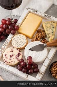 Glass of red wine with selection of various cheese in wooden box and grapes on light background. Blue Stilton, Red Leicester and Brie Cheese with Cheddar and knife.