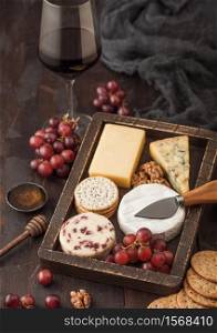 Glass of red wine with selection of various cheese in wooden box and grapes on wooden background. Blue Stilton, Red Leicester and Brie Cheese and bowl of nuts with honey.