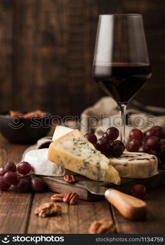 Glass of red wine with selection of various cheese on the board and grapes on wooden table background. Blue Stilton, Red Leicester and Brie Cheese.