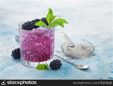 Glass of refreshing summer cocktail with blackberry, ice and mint on blue background with bar spoon and strainer.