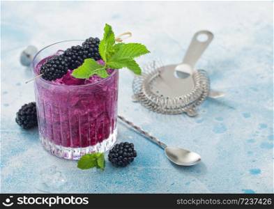 Glass of refreshing summer cocktail with blackberry, ice and mint on blue background with bar spoon and strainer. Top view