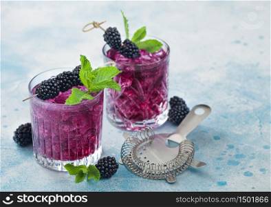 Glass of refreshing summer cocktail with blackberry with ice cubes and mint on light blue background with strainer. Soda and alcohol mix.