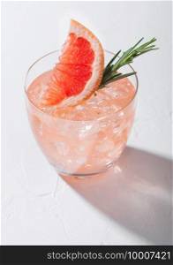 Glass of refreshing summer red grapefruit cocktail with ice cubes, fruit slice and rosemary on white background with deep shadow. Hard light