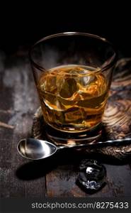 Glass of single malt whiskey with bar spoon and ice cubes on wooden background.Macro.