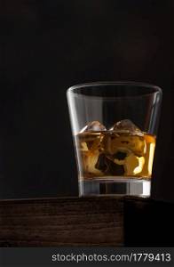 Glass of single malt whiskey with ice cubaes on top of wooden box on black background