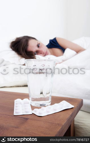 Glass of water and two strips of pain killers on the bed side table with a young, sick, woman in the background