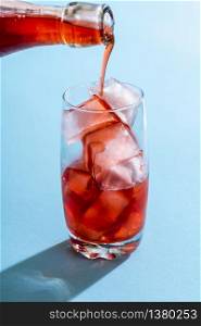 Glass of water with ice cubes and strawberry syrup. Pouring syrup from a glass bottle to a glass full of ice cubes. Drink preparation in bright light on a blue colored background