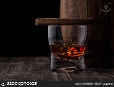 Glass of whiskey with ice cubes and cigar next wooden barrel. Cognac and brandy drink