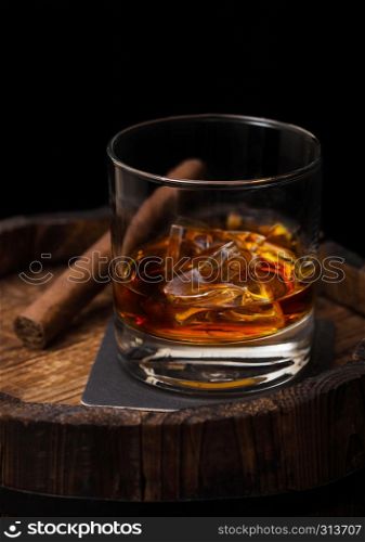 Glass of whiskey with ice cubes and cigar on top of wooden barrel. Cognac and brandy drink