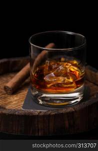 Glass of whiskey with ice cubes and cigar on top of wooden barrel. Cognac and brandy drink