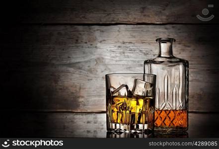 Glass of whiskey with ice cubes and faceted bottle on a wooden background