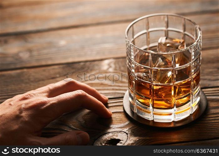 Glass of whiskey with ice cubes and man&rsquo;s hand on rustic wooden table with copy-space. Alcoholism concept.